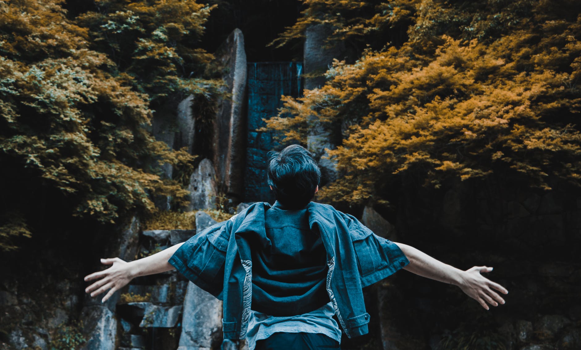 person spreading his hands in front of gray concrete structure surrounded by brown and green leaf trees